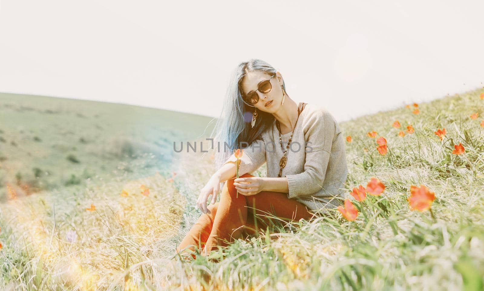 Beautiful young woman in sunglasses sitting on red tulip meadow on sunny day, looking at camera.