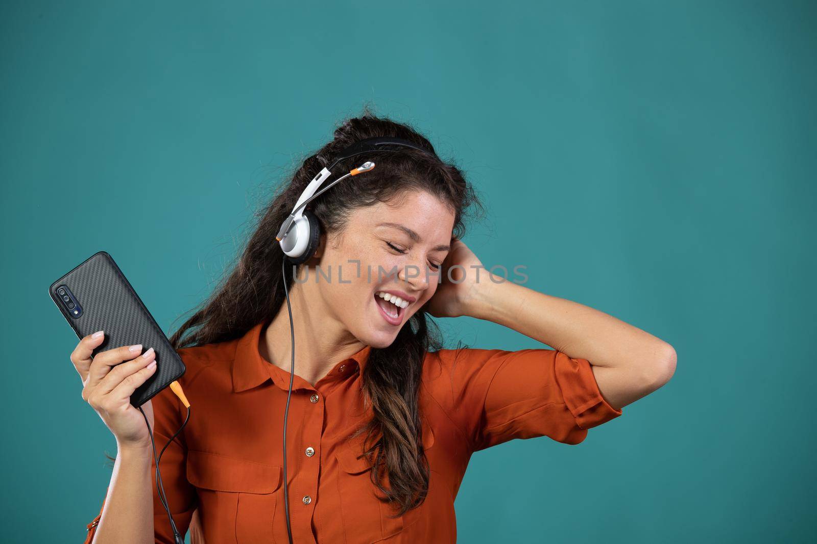 Pretty young woman listening music with headphones, holding mobile phone and singing, on blue background in studio
