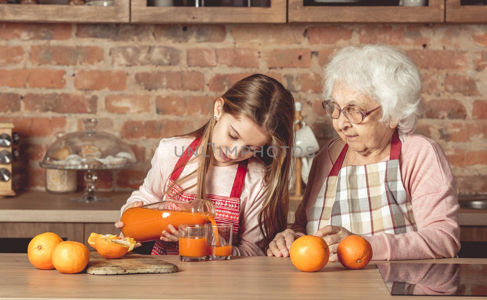 Little granddaughter making fresh orange juice with her senior grandmother on rustic kitchen