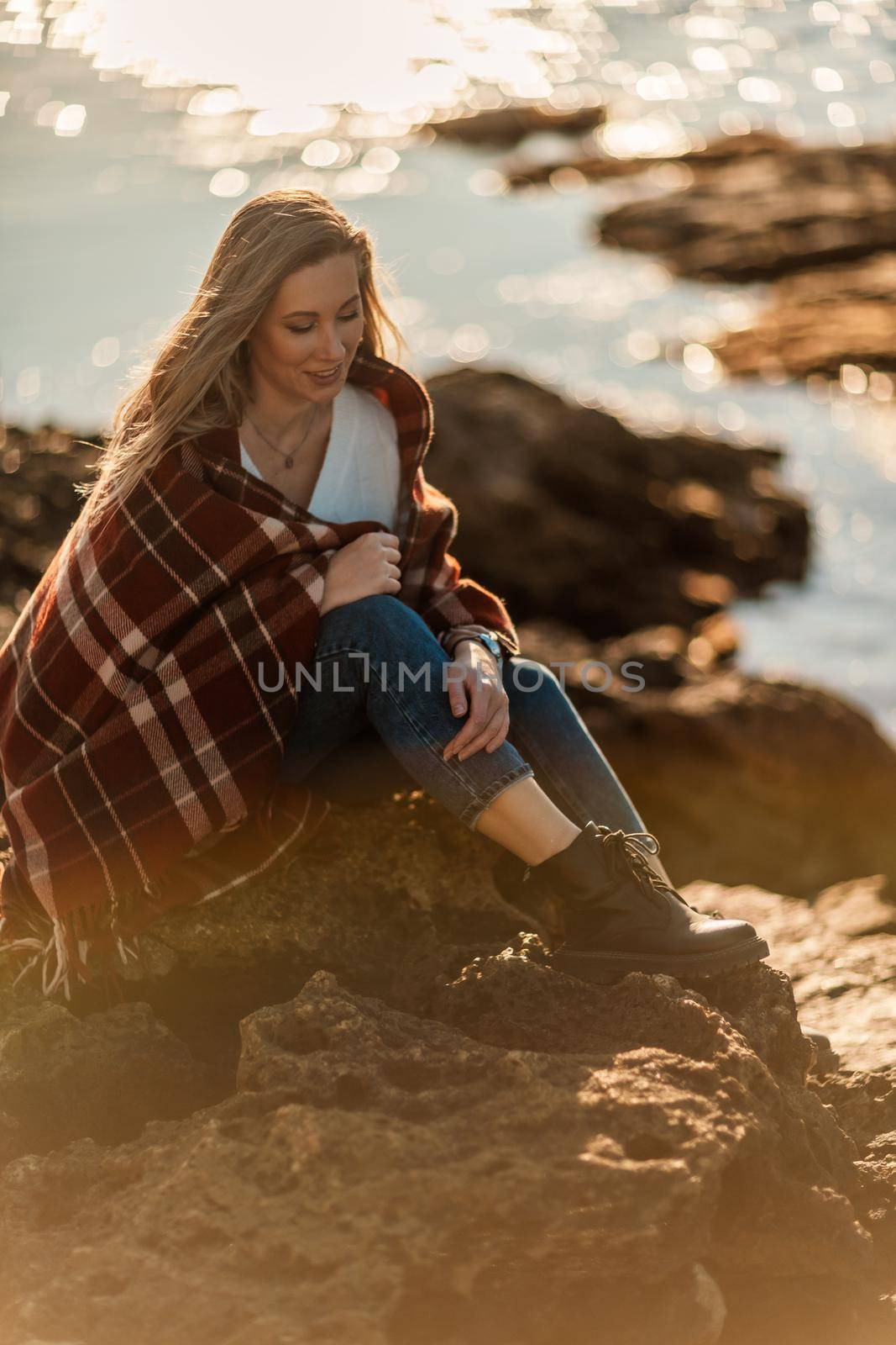 Attractive blonde Caucasian woman enjoying time on the beach at sunset, sitting in a blanket and looking to the side, with the sunset sky and sea in the background. Beach vacation