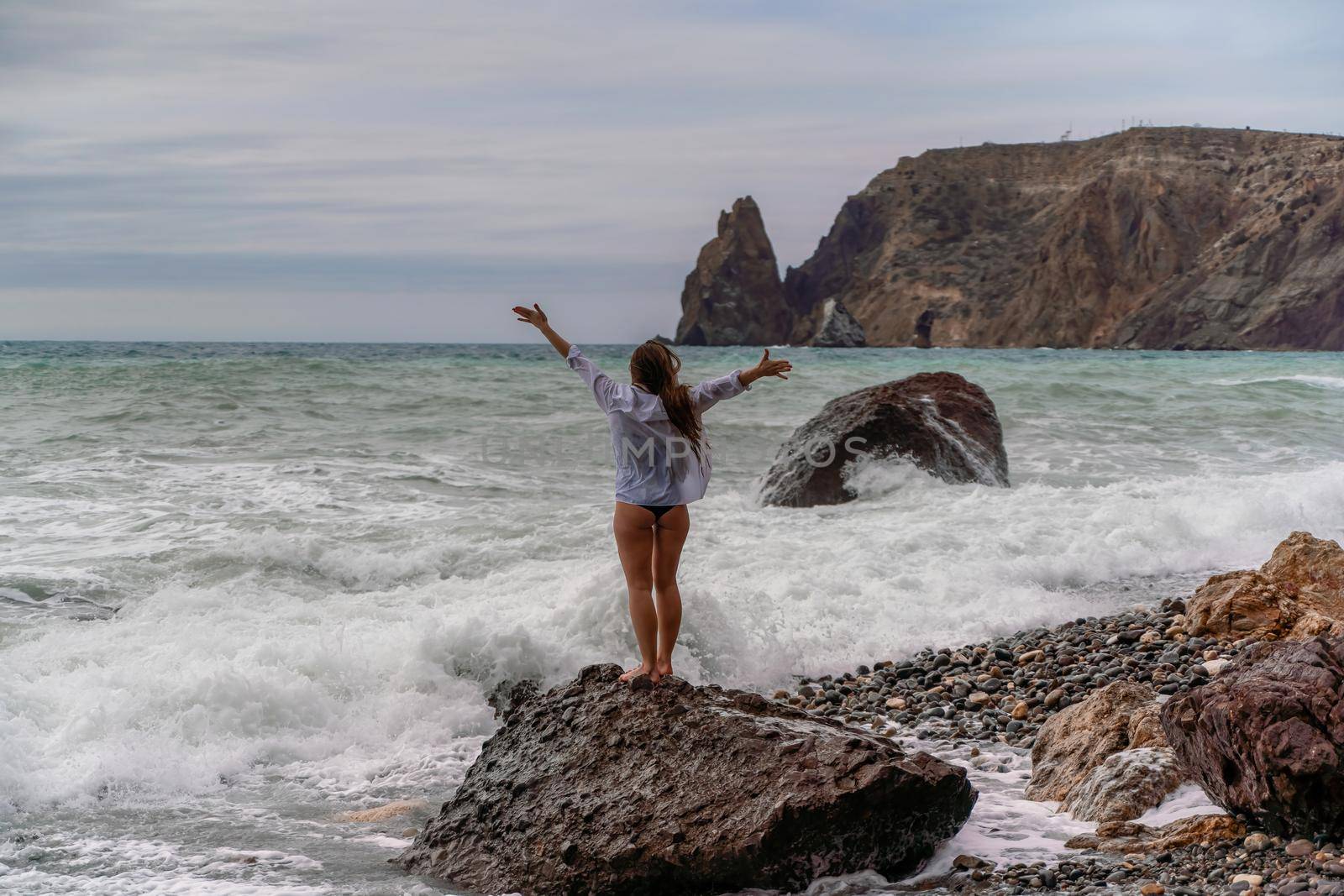 A beautiful girl in a white shirt and black swimsuit stands on a rock, big waves with white foam. A cloudy stormy day at sea, with clouds and big waves hitting the rocks