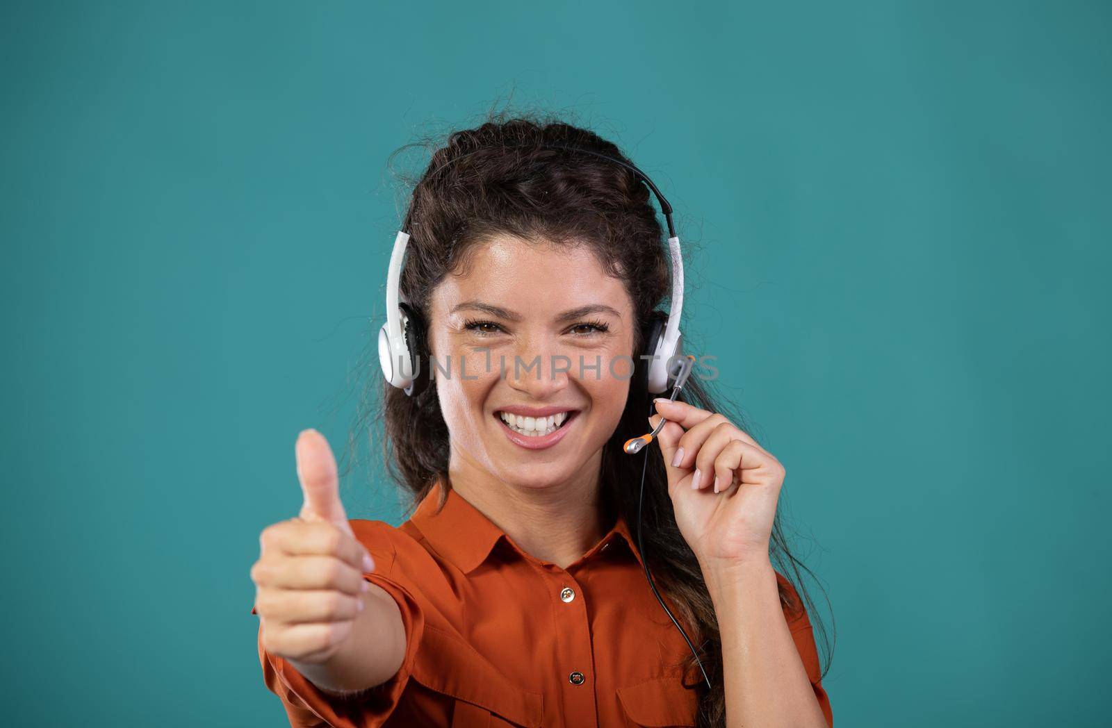 Satisfied pretty woman operator with headphones on head showing thumb up gesture in studio on blue background