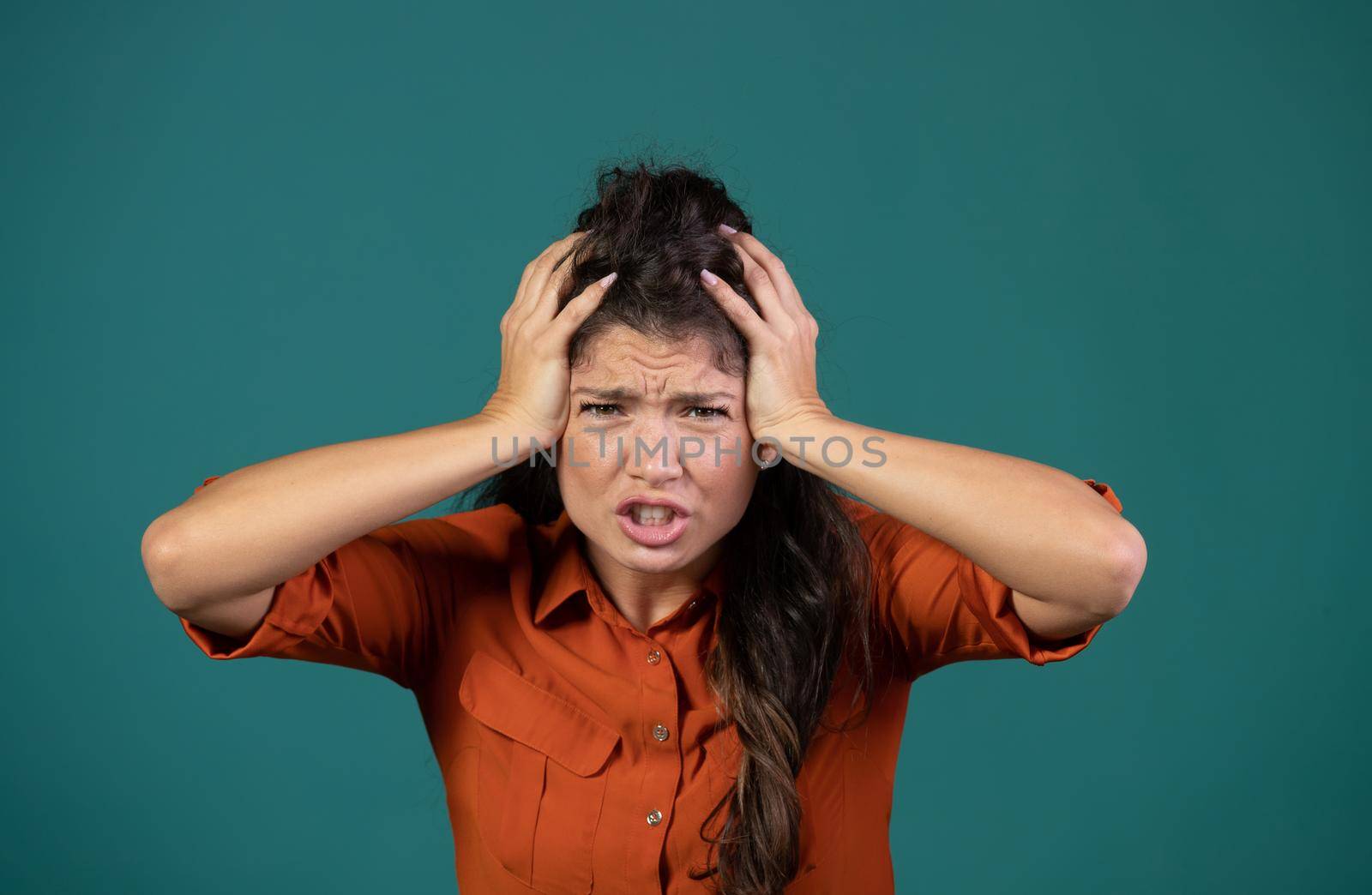 Frustrated young woman holding hands on head, madness concept, in studio on blue background