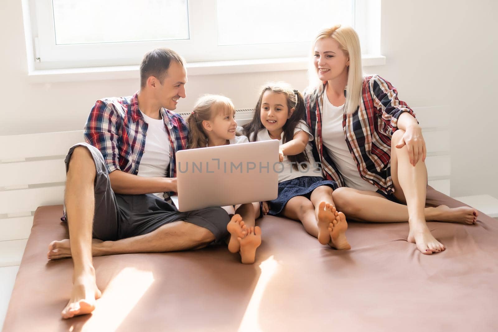 Happy family concept. Beautiful mother and handsome father with their daughters spending time together at home and lying on bed with laptop.