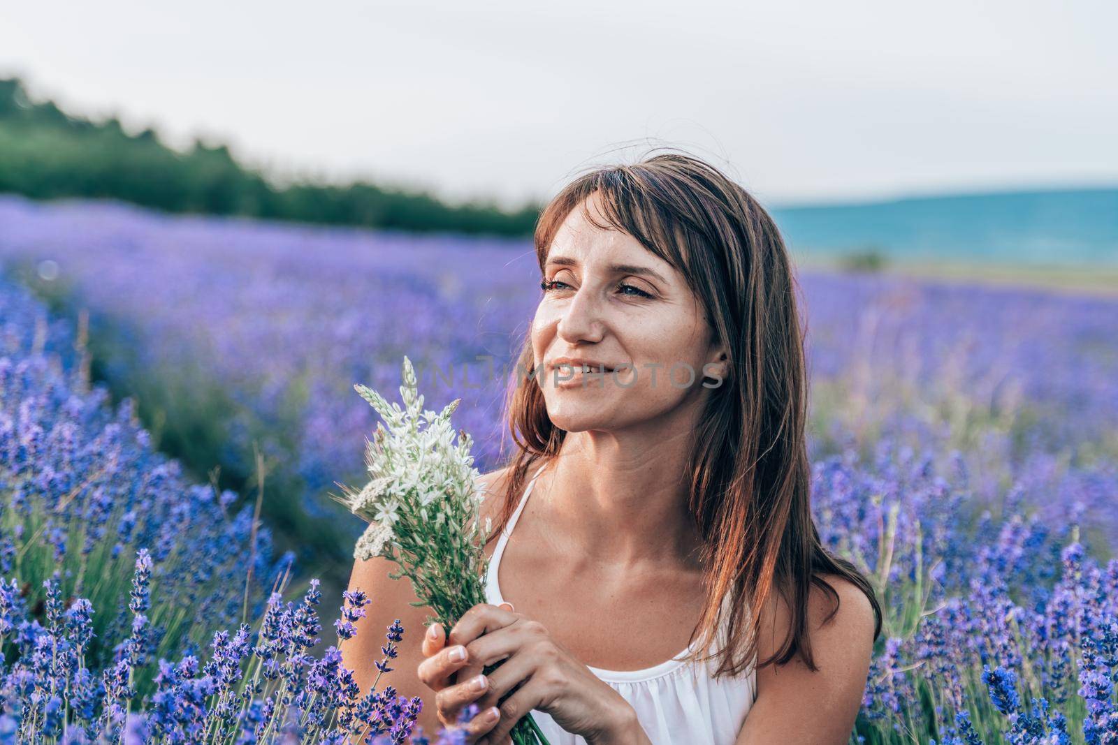 Close up portrait of happy young brunette woman in white dress on blooming fragrant lavender fields with endless rows. Warm sunset light. Bushes of lavender purple aromatic flowers on lavender fields. by panophotograph