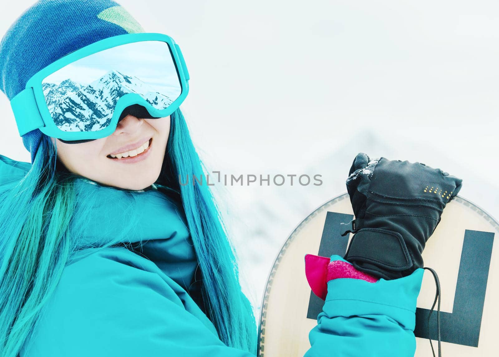 Smiling young woman with blue hair in protective sunglasses standing with snowboard, looking at camera. Snow mountains reflected in glasses.