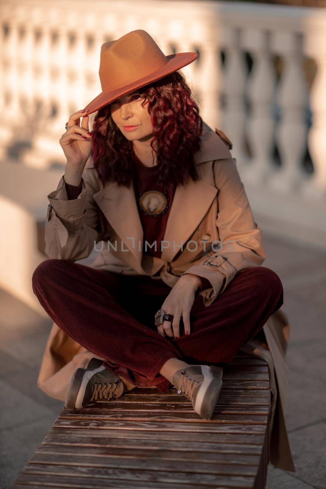 Outdoor fashion portrait of young elegant fashionable brunette woman, model in stylish hat, choker and light raincoat posing at sunset in European city. by Matiunina