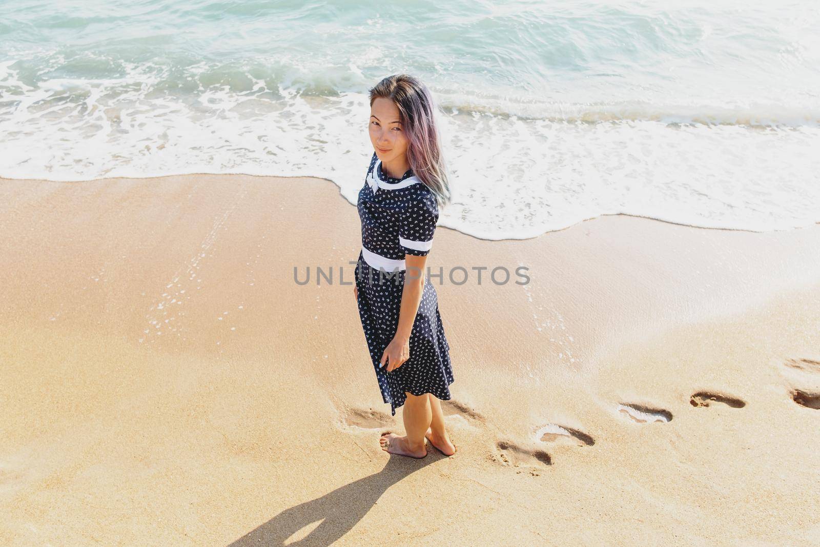 Beautiful barefoot young woman in dress walking on sand beach near the sea, looking at camera.