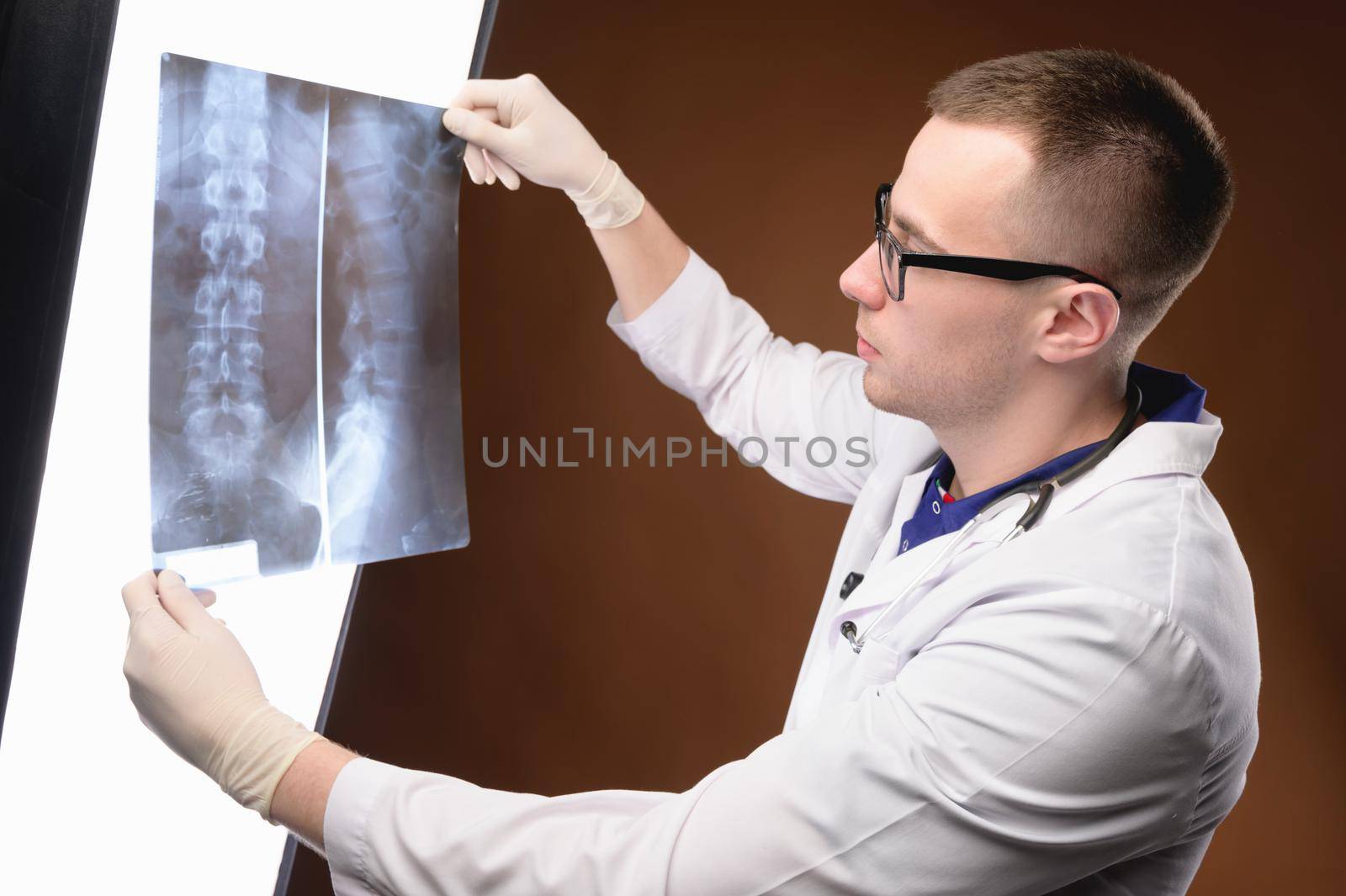Young doctor radiologist in glasses and medical clothes looks at the lumen x-ray picture of the spine on a brown background
