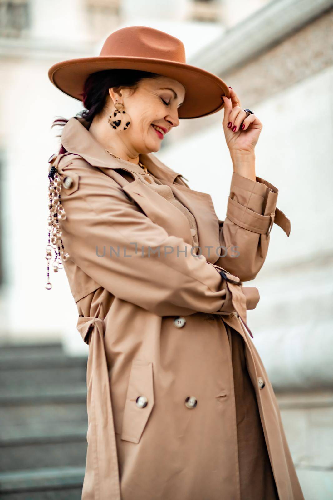 Outdoor fashion portrait of young elegant fashionable brunette woman, model in stylish hat, choker and light raincoat posing at sunset in European city. by Matiunina