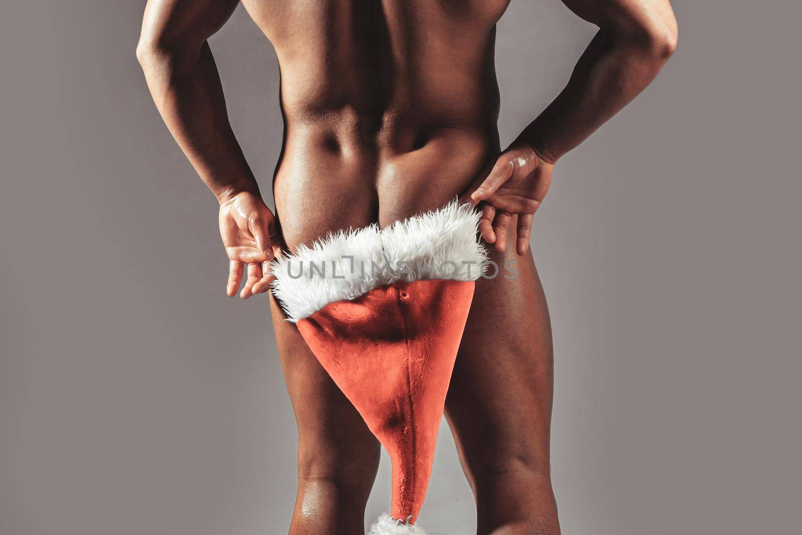 Sexy muscular gay man hiding buttocks butt, ass under red santa claus hat. Winter holidays celebration on grey.