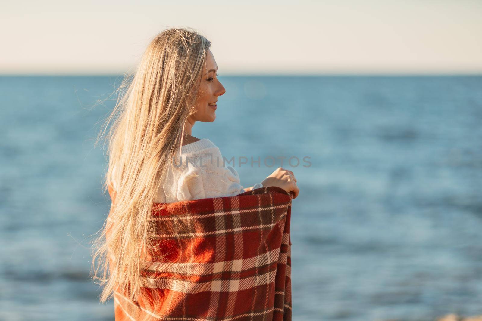 Attractive blonde Caucasian woman enjoying time on the beach at sunset, walking in a blanket and looking to the side, with the sunset sky and sea in the background. Beach vacation.