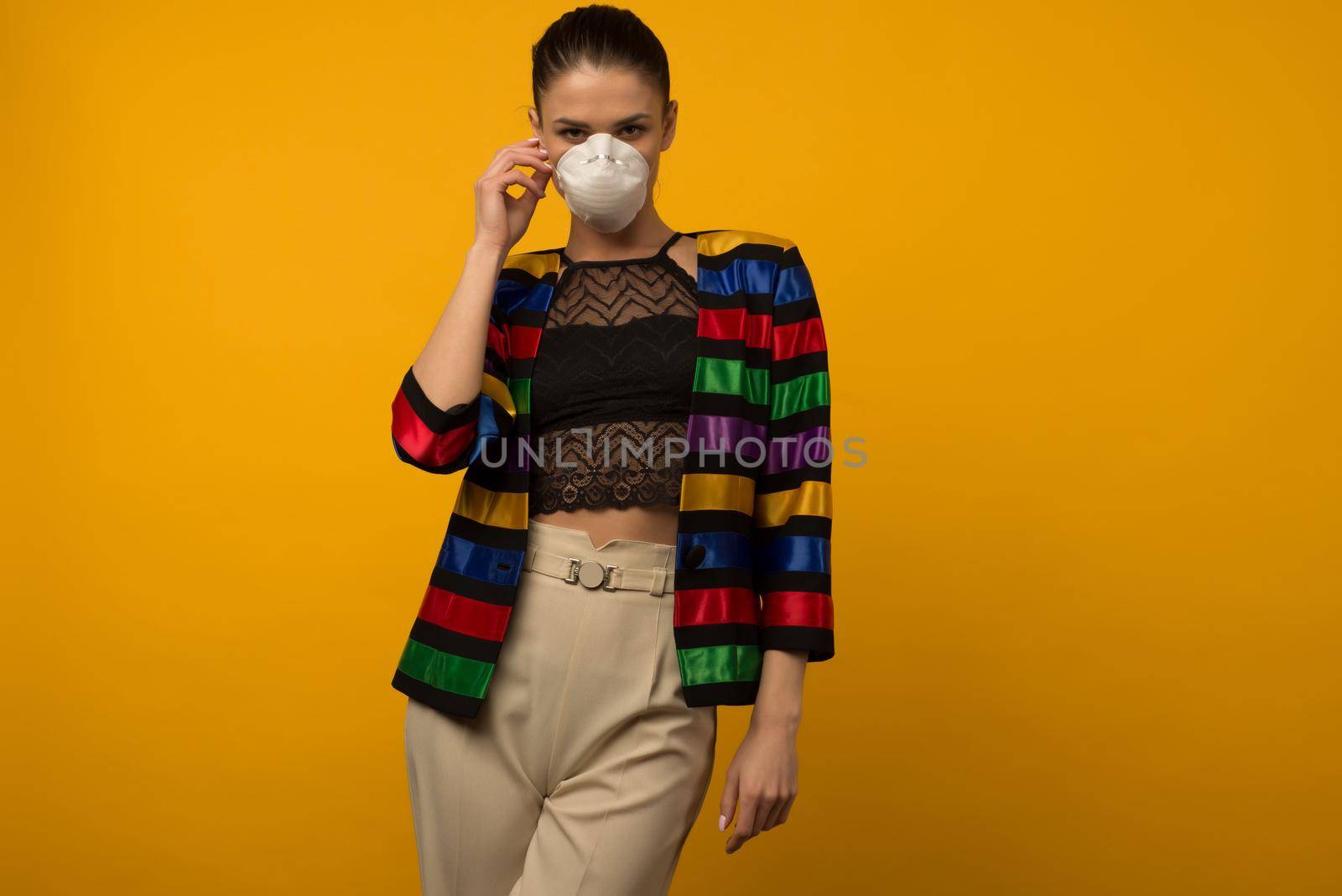 Beautiful slim girl fashion model posing in a protective respirator on a yellow background. LGBT community rainbow color jacket