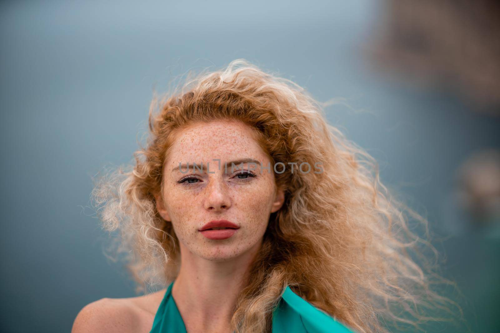 Outdoor portrait of a young beautiful natural redhead girl with freckles, long curly hair, in an emerald dress, posing against the background of the sea