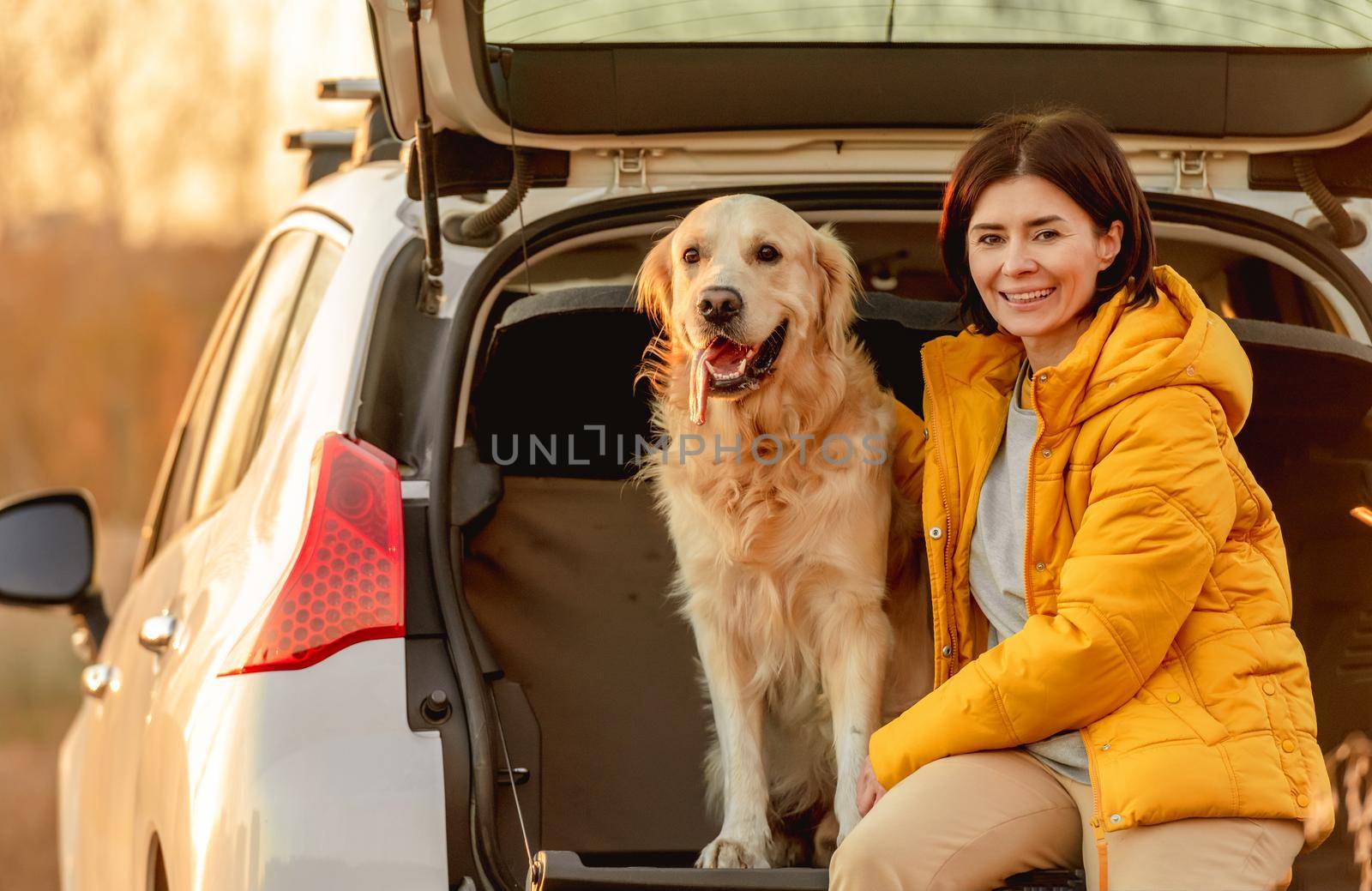 Girl with golden retriever dog sitting in car trunk outdoors. Young woman with doggy pet labrador in vehicle at nature at autumn