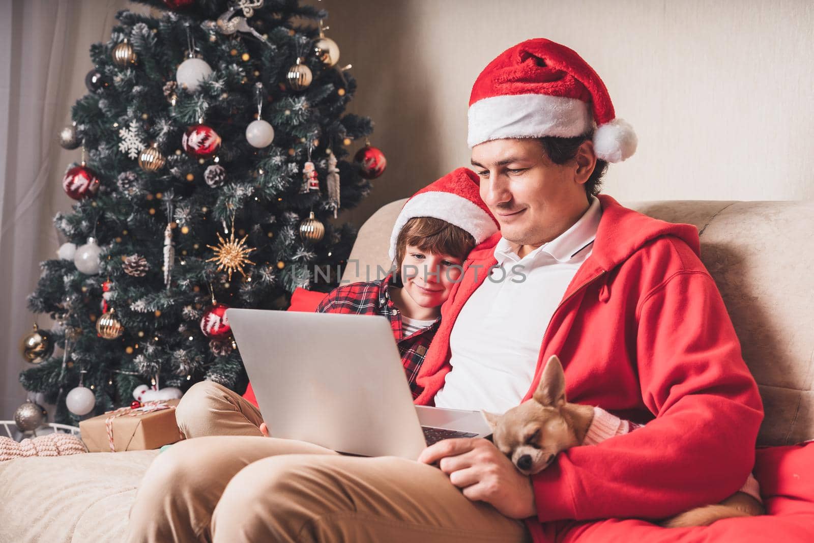 Father with child and puppy dog in Santa hats having a video call on Christmas day at laptop, sitting on a couch in the living room with Christmas tree at home. by Ostanina
