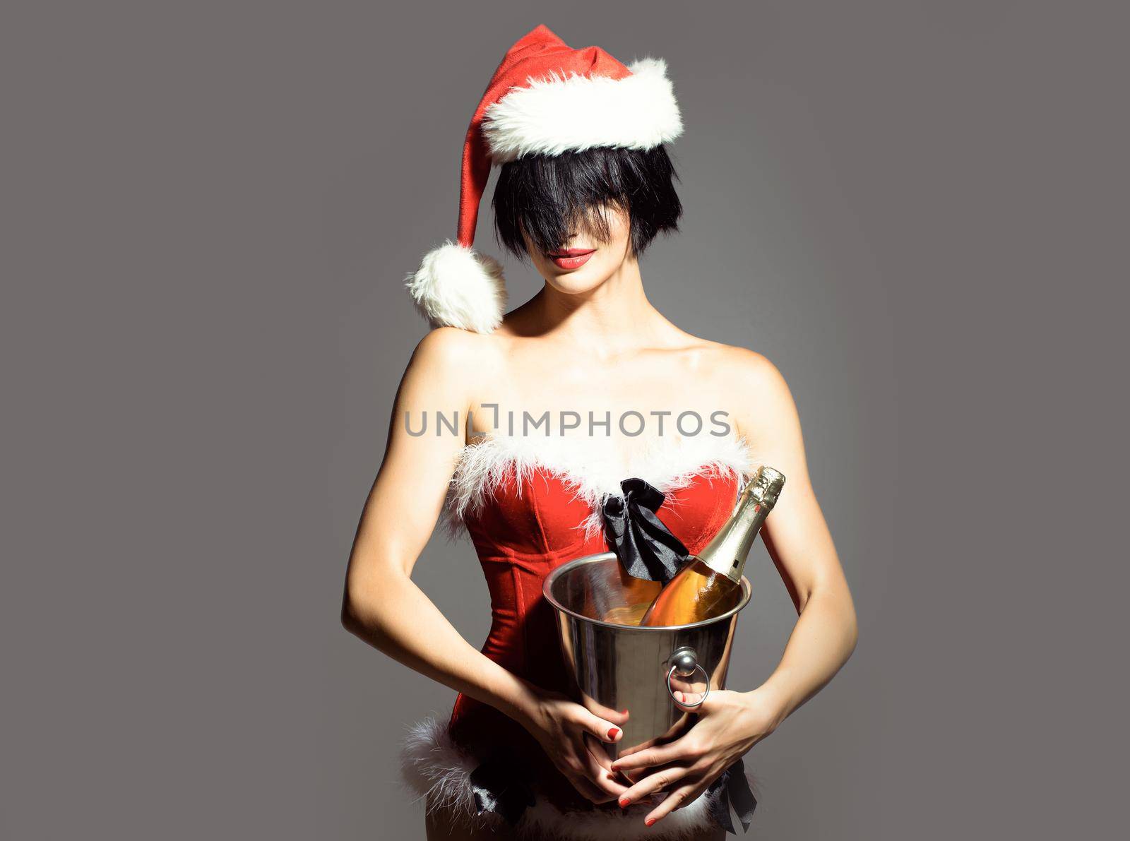 Sexy woman or santa girl with red lips on pretty face in Christmas hat and holiday costume holds pail with wine or champagne bottle, on grey background. by Tverdokhlib