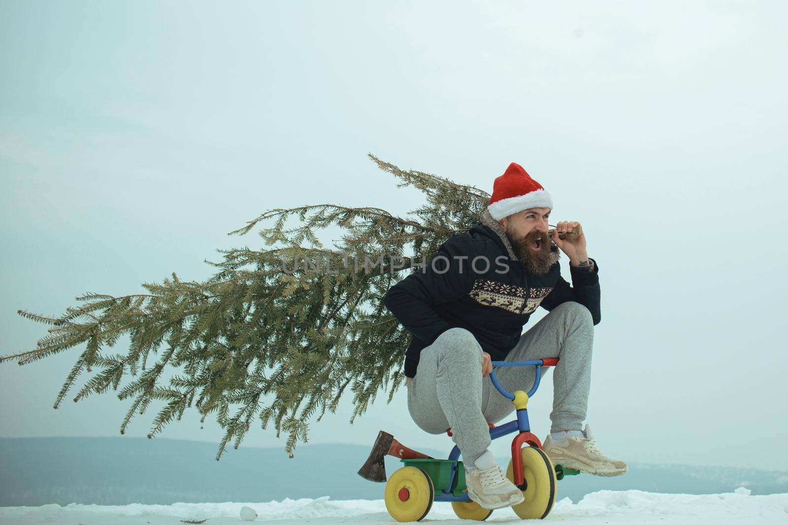 Hipster carrying Christmas tree on white snow. Excited crazy funny cyclist in santa hat celebration Christmas and new year. Man riding bicycle tricycle on snowy landscape by Tverdokhlib
