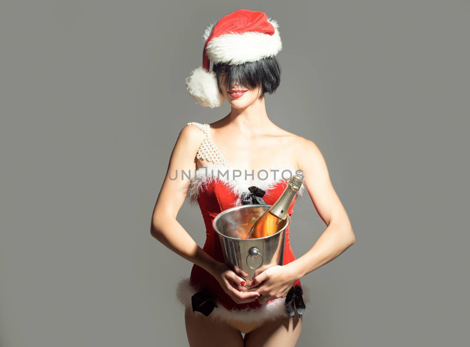 Sexy young woman or santa girl in Christmas hat and holiday costume holds metallic pail with wine or champagne bottle on grey background.