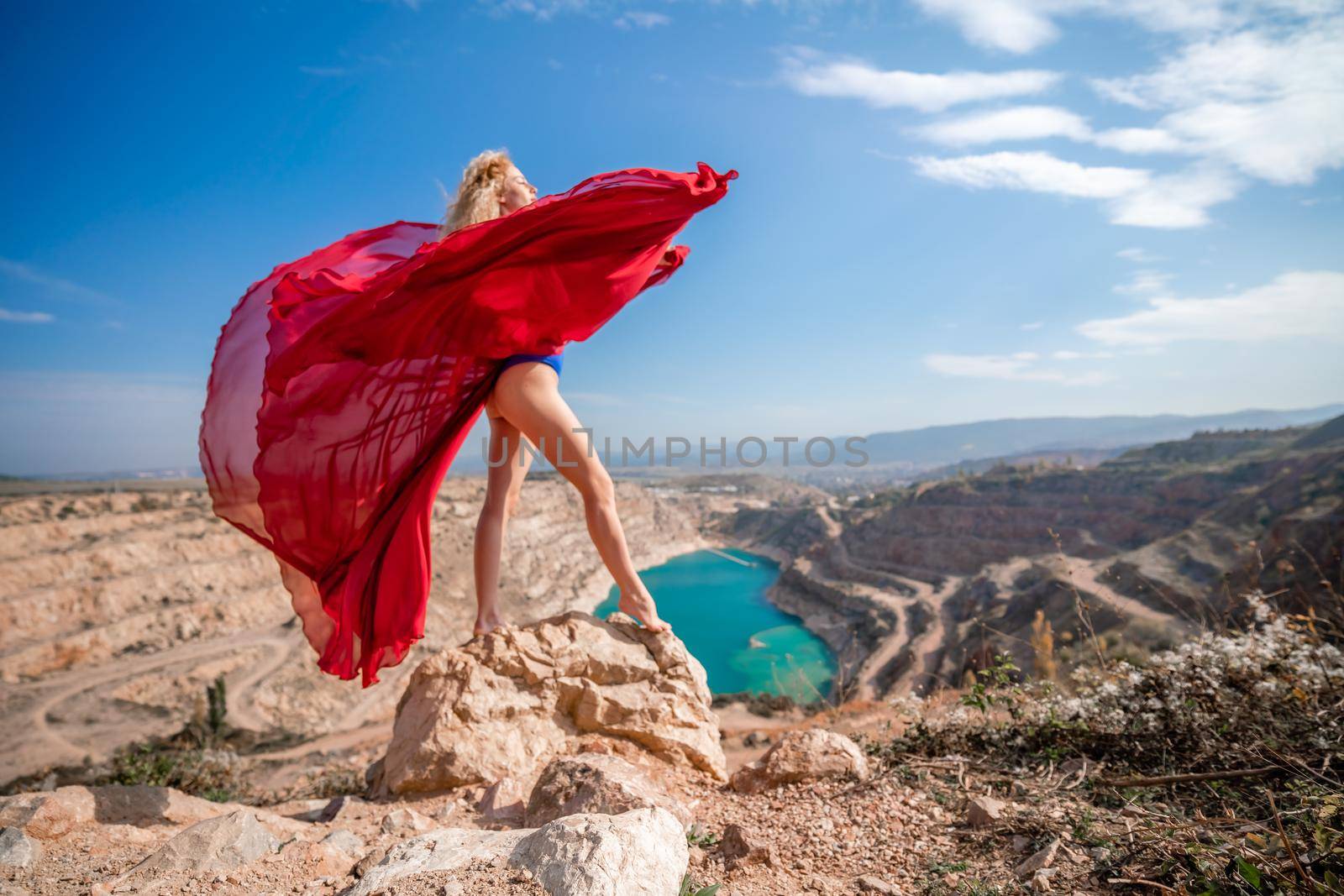 Side view of a beautiful sensual woman in a red long dress posing on a rock high above the lake in the afternoon. Against the background of the blue sky and the lake in the form of a heart by Matiunina