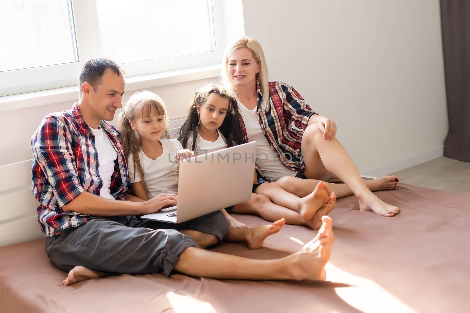 Happy family concept. Beautiful mother and handsome father with their daughters spending time together at home and lying on bed with laptop.