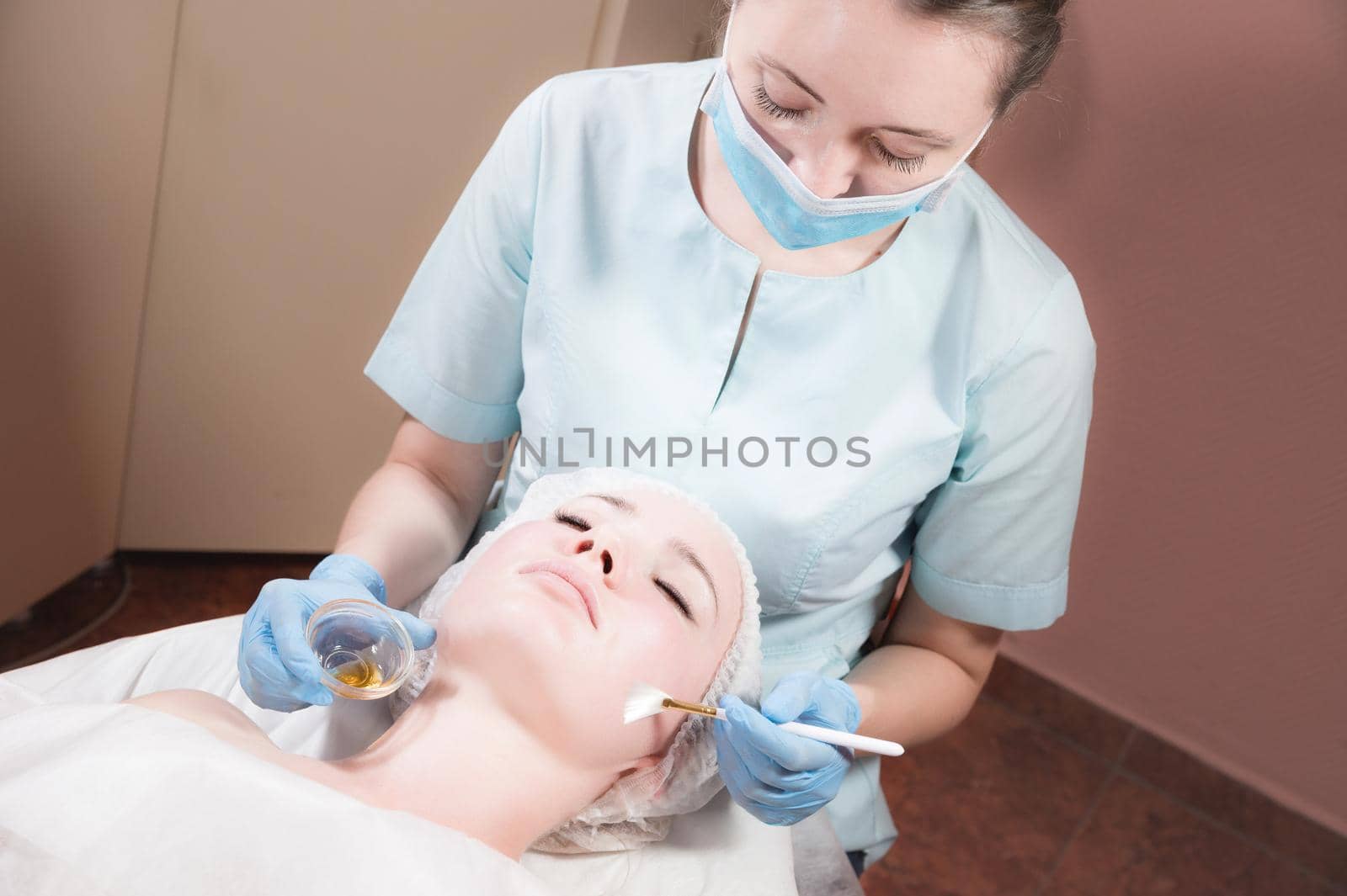 face of a young woman close-up lying down cosmetic procedure is performed applying a rejuvenating mask. by a professional with a brush in blue gloves