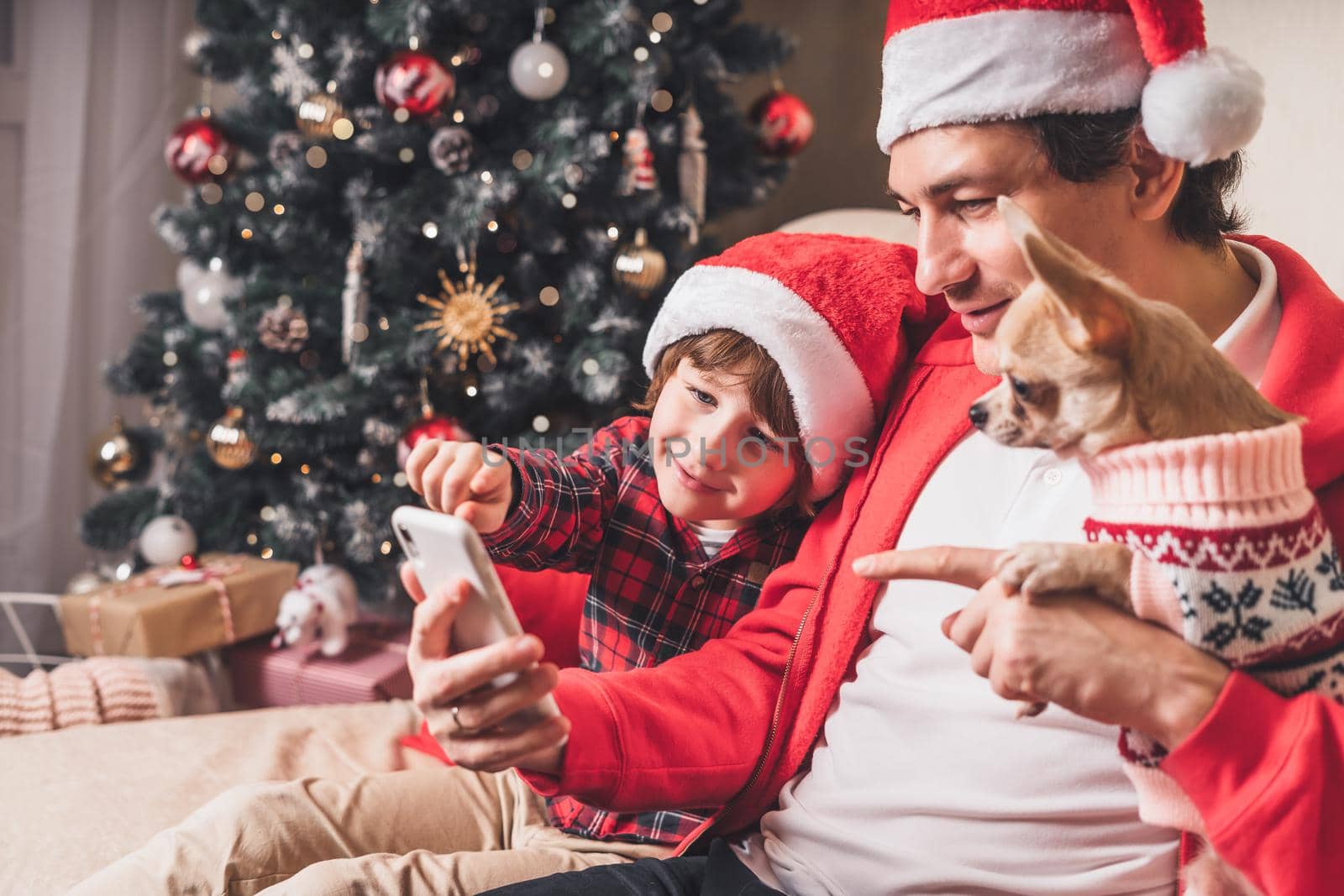 Child with Father and puppy dog in Santa hats having a video call on Christmas day, sitting on a couch in the living room with Christmas tree at home. by Ostanina