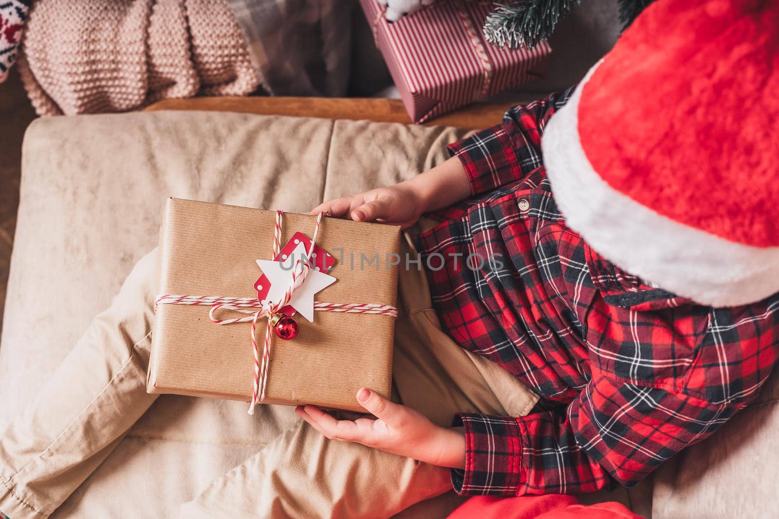Close-up of child hands holding and open Christmas gift at winter holidays. Kid boy at home sitting on couch, top view.