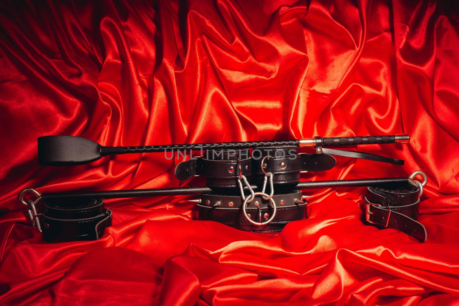 Close up bdsm outfit. Bondage, kinky adult sex games, kink and BDSM lifestyle concept with a pair of leather handcuffs, flogger, whip on red silk with copy space.