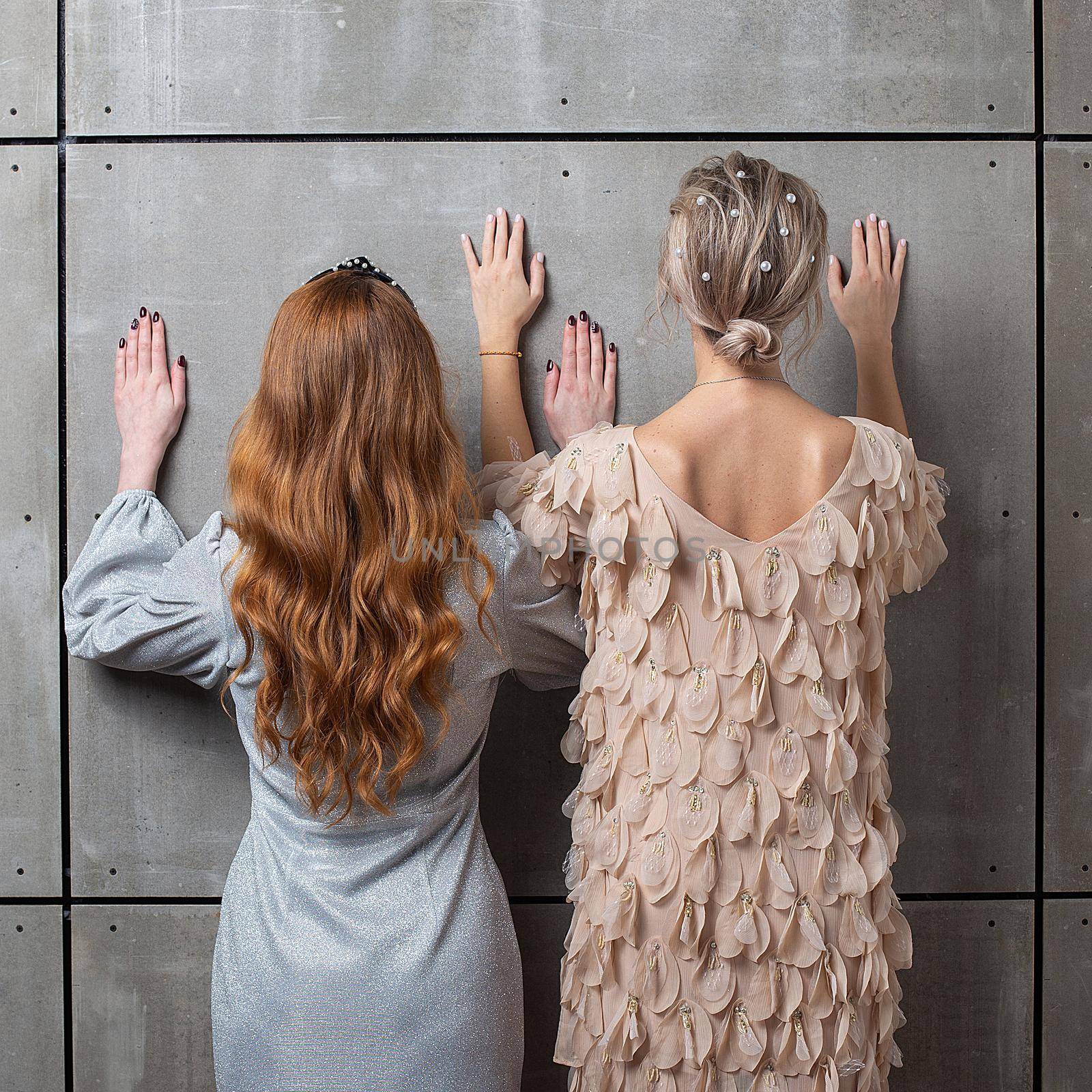 two girls standing against gray concrete wall with raised hands, rear view. females lean their hands on wall. ladies stand with hands up in beautiful dresses with gorgeous haircuts. fashion shoot