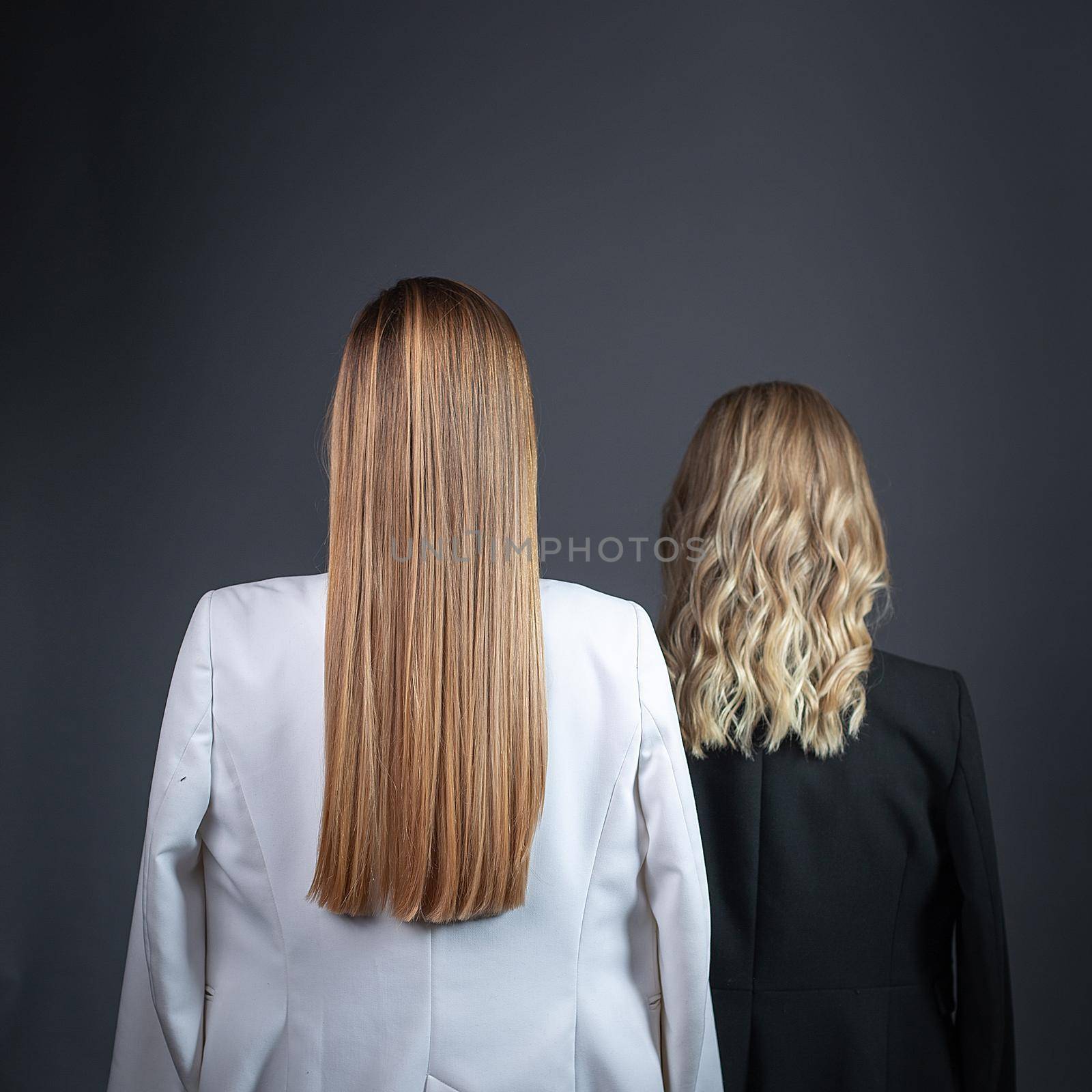 two girls with long blond hair standing against gray background in strict suits, rear view. females turned around at wall. ladies stand with their backs turned to camera. studio photoshoot