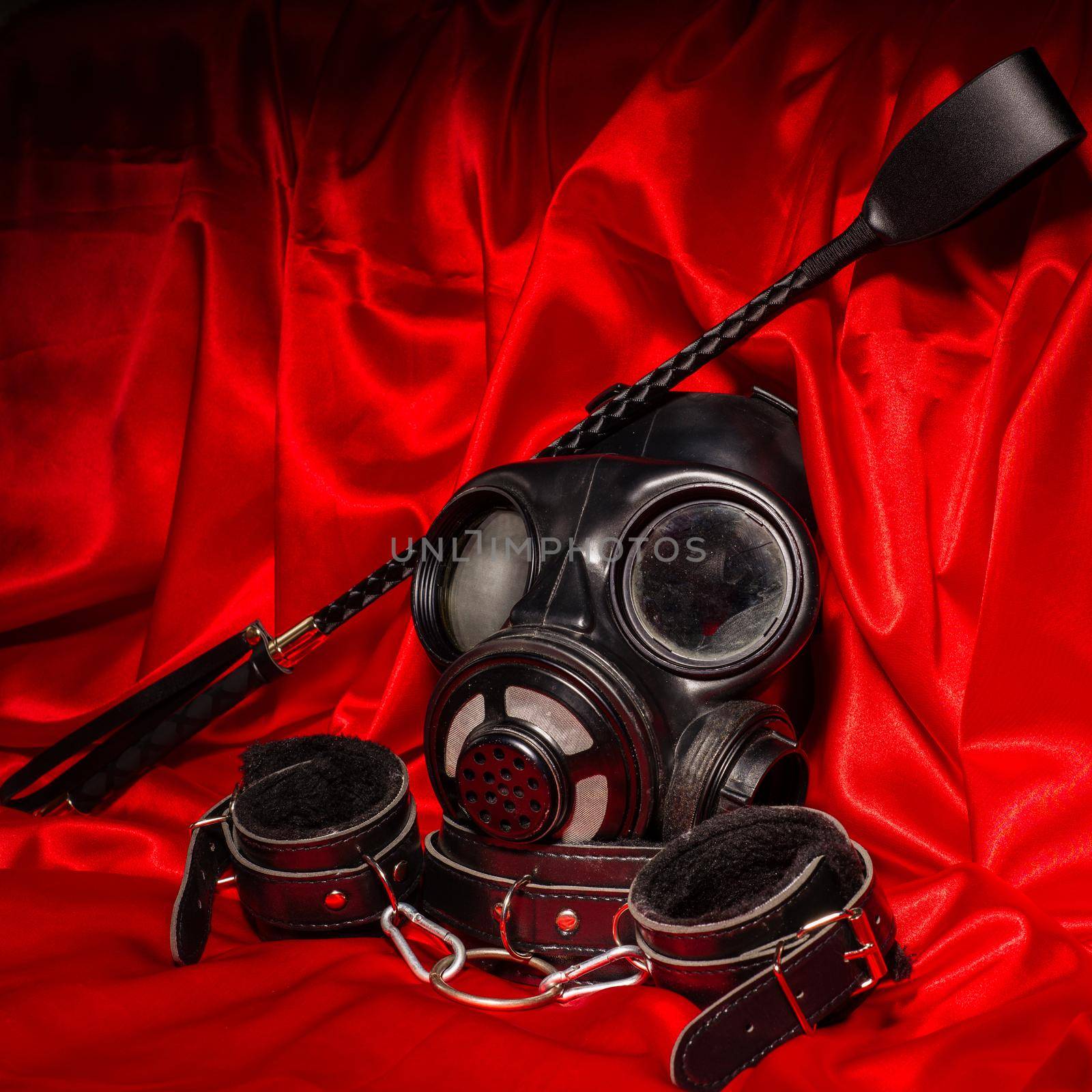 Close up bdsm outfit. Bondage, kinky adult sex games, kink and BDSM lifestyle concept with gas mask, whip, collar, leather handcuffs on red silk with copy space.