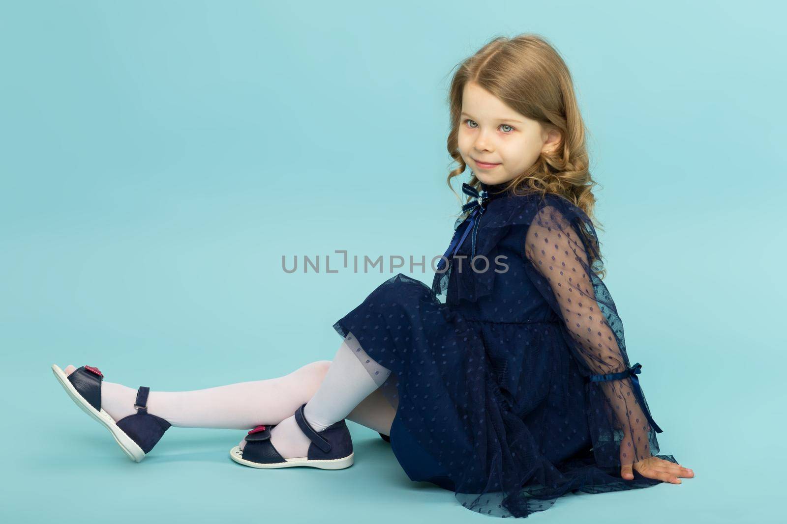 Happy little girl in blue dress sitting on floor. Portrait of adorable blonde girl wearing beautiful lace dress sitting sideways to camera on light blue background in studio.