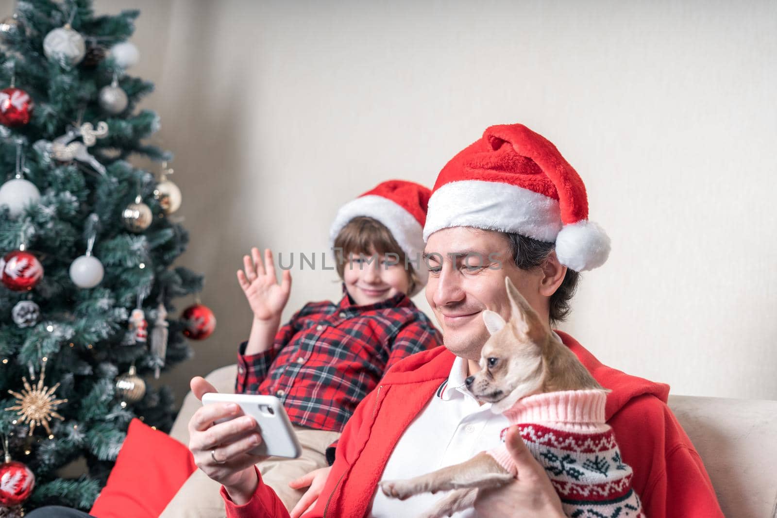 Father with child and puppy dog in Santa hats having a video call on Christmas day at smartphone, sitting on a couch in the living room with Christmas tree at home.