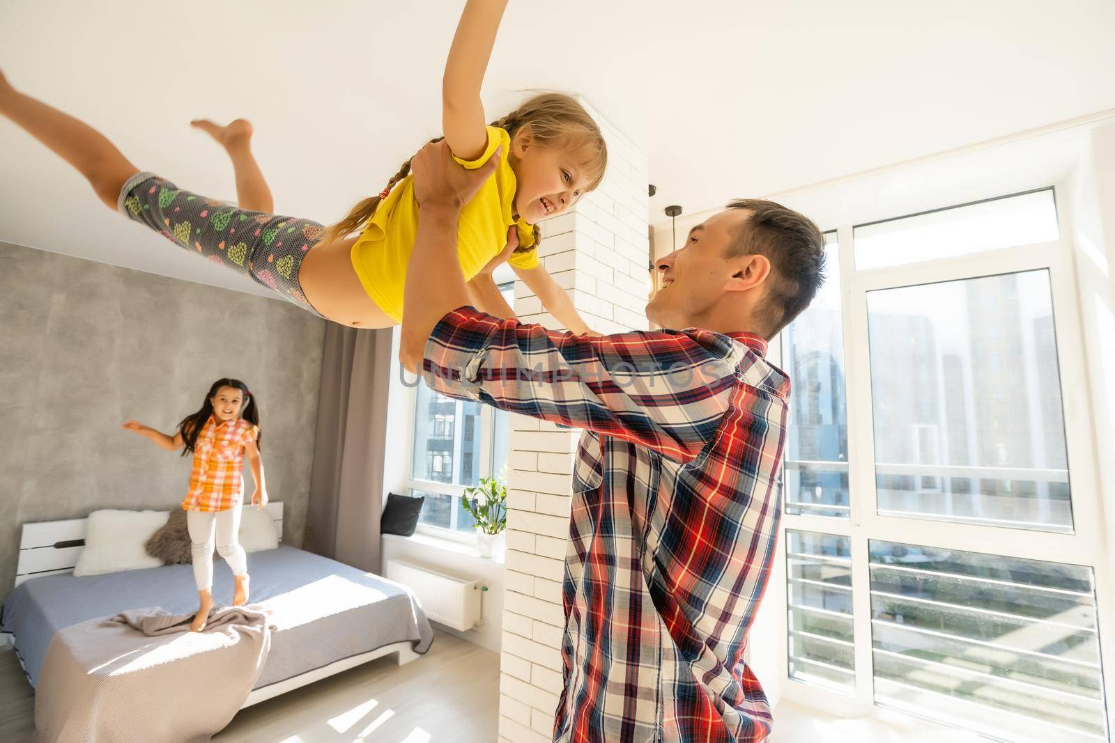 Overjoyed young father holding two little children siblings, having fun together at home, head shot. Excited dad playing with joyful adorable small daughter sisters, enjoying tender weekend moment.