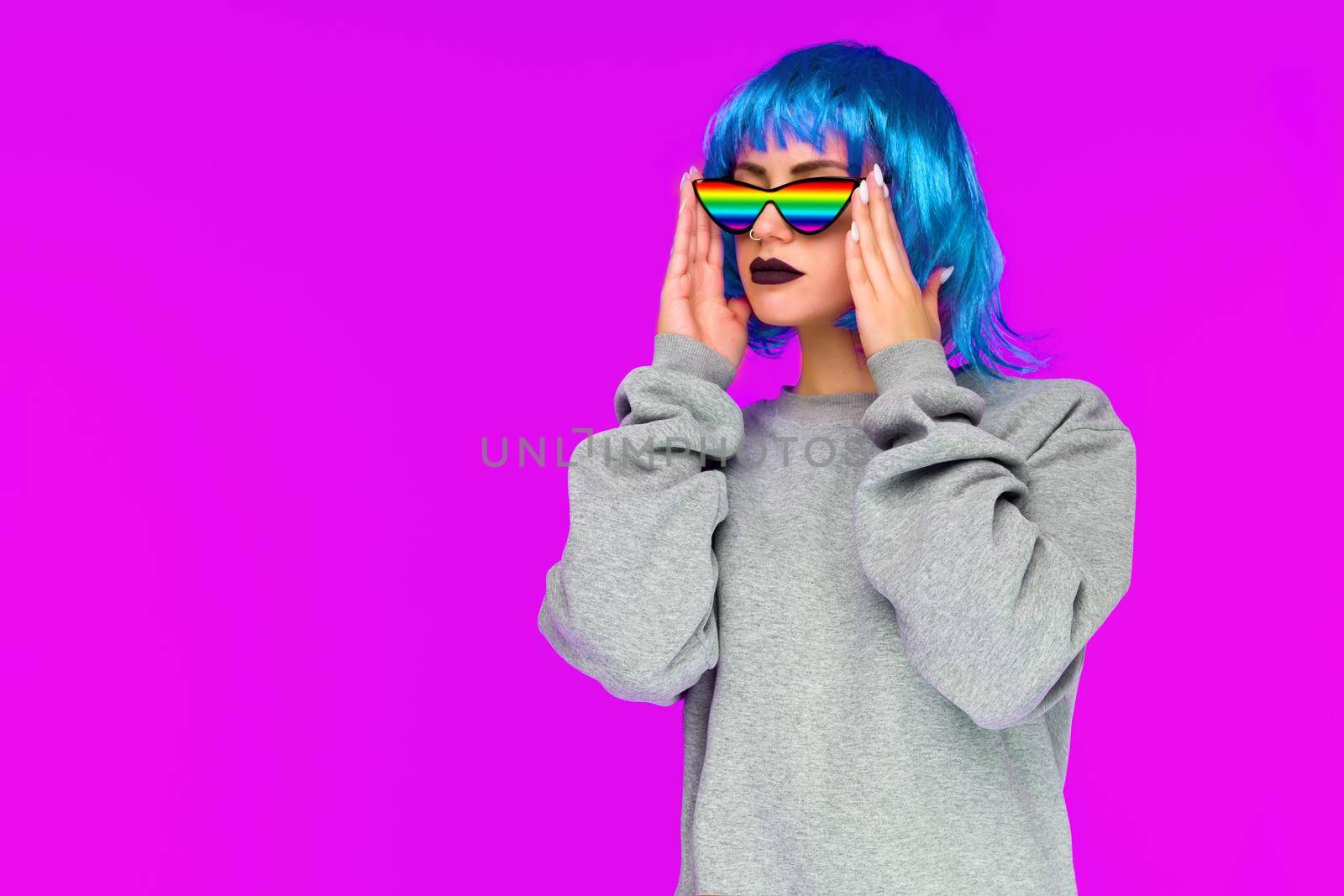 Lesbian girl in blue wig and rainbow sunglasses looks to the side on pink background - image