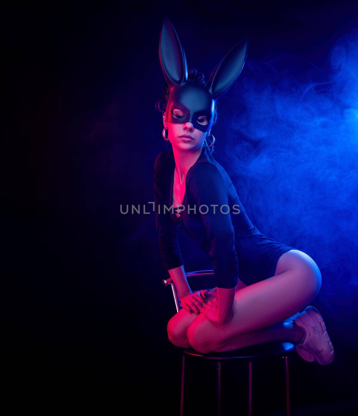 the beautiful girl in a bodysuit poses in a photo Studio on a dark background in neon light. On the face of a rabbit mask