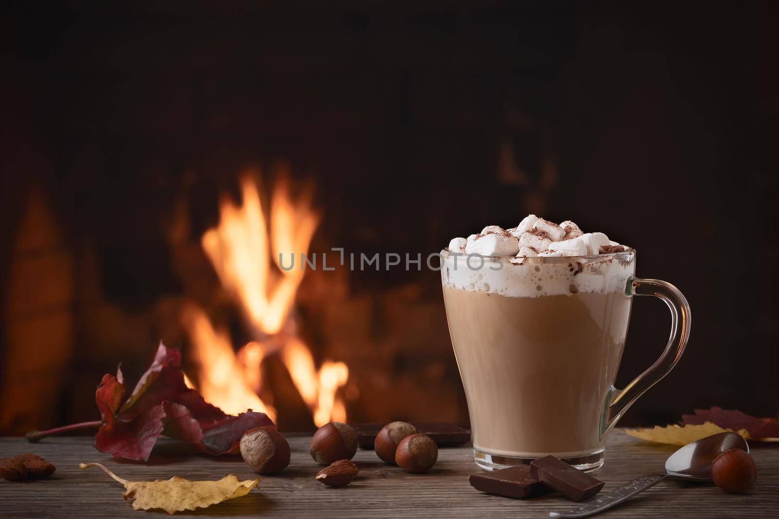 Cocoa with marshmallows and chocolate in a glass mug on a wooden table near a burning fireplace by galsand