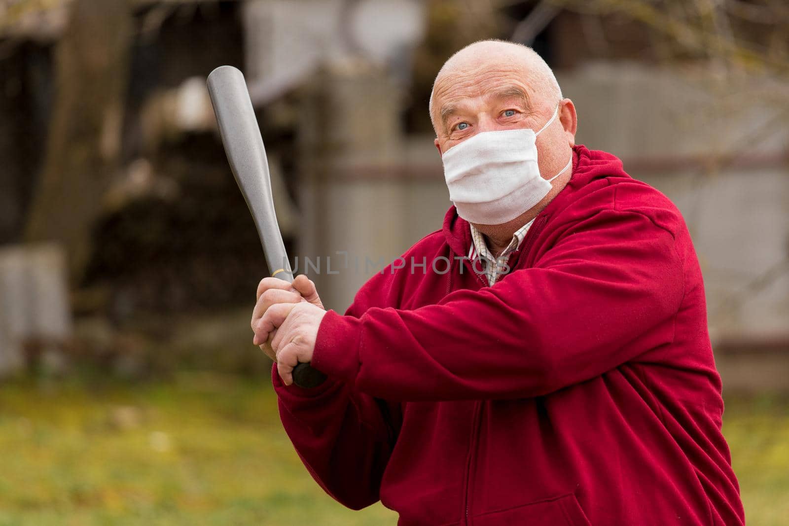 Angry aggressive elderly man in protective safe medical mask swings baseball bat in the background of outdoor street, portrait, close up.