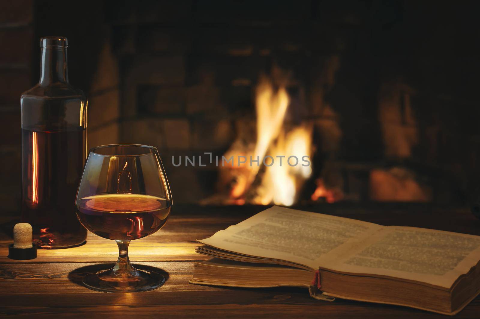 Glass of cognac, a bottle and an open old book on the table near the burning fireplace. Rest and relaxation concept by galsand