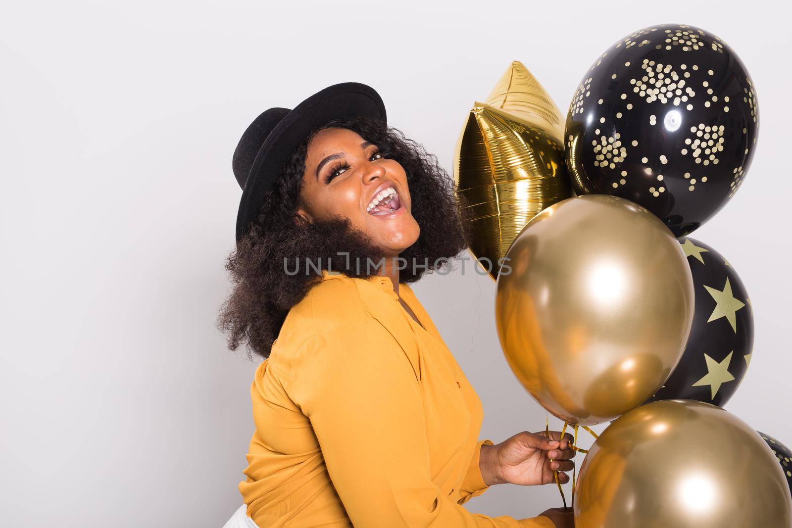 Portrait of smiling young African-American adult woman looking sweet on yellow background holding balloons.