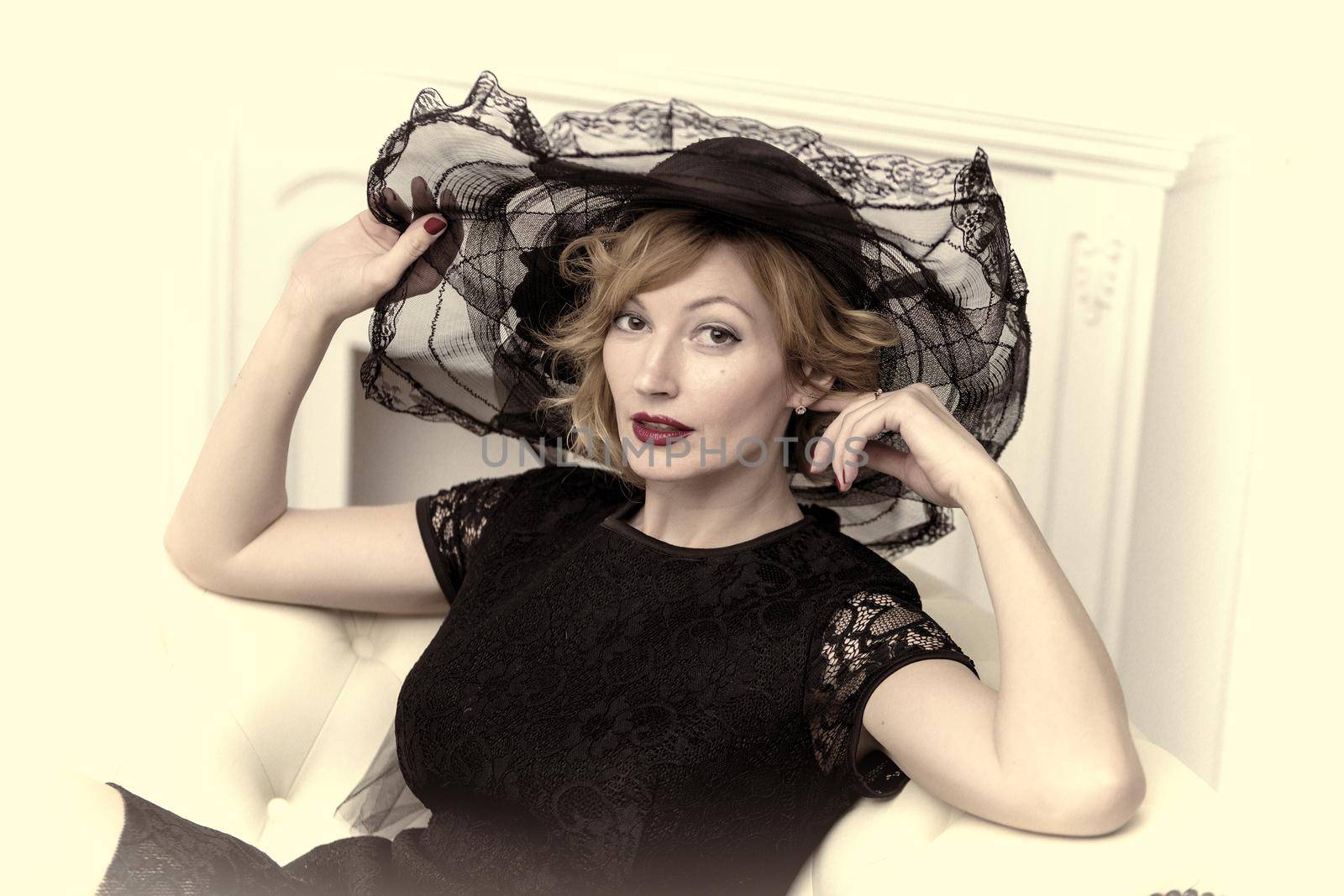 A beautiful young woman in a luxurious hat sits on a white leather sofa near the fireplace. Retro style