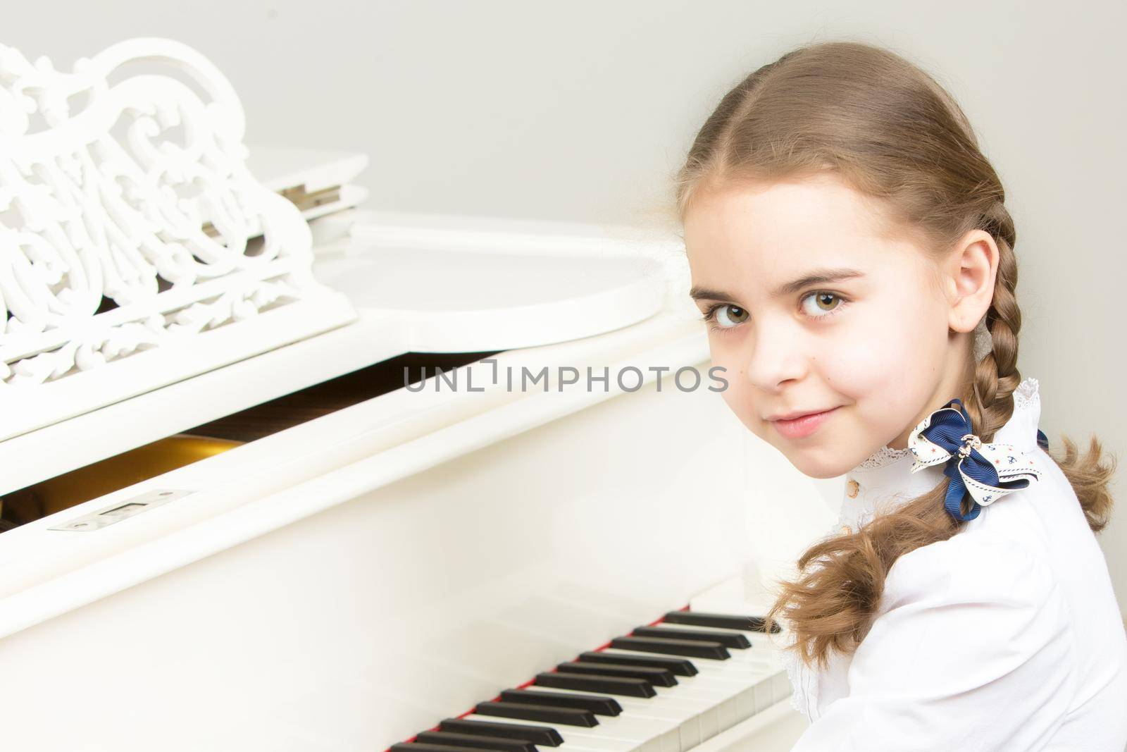 Charming little blonde with long wattled hair in plaits, playing on a white grand piano. In musical school.