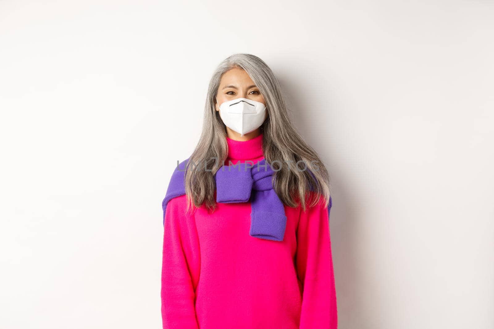 Covid, pandemic and social distancing concept. Cheerful asian senior female model in face mask smiling at camera, preventive measures from coronavirus, white background.