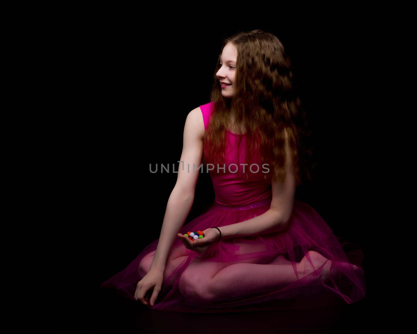 A beautiful little gymnast girl of school age holds multicolored confetti in her palms. Studio photography on a black background.