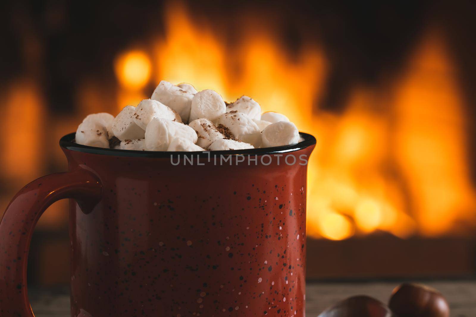 Cocoa with marshmallows and chocolate in a red mug on a wooden table near a burning fireplace.