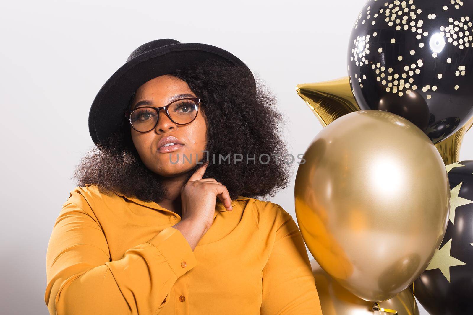 Portrait of smiling young African-American adult woman looking sweet on yellow background holding balloons.