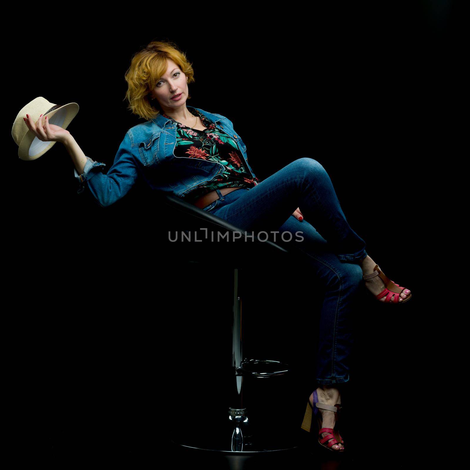Beautiful young woman posing in a studio studio on a bar stool holding a hat in her hand. The concept of style and fashion. Isolated on black background.