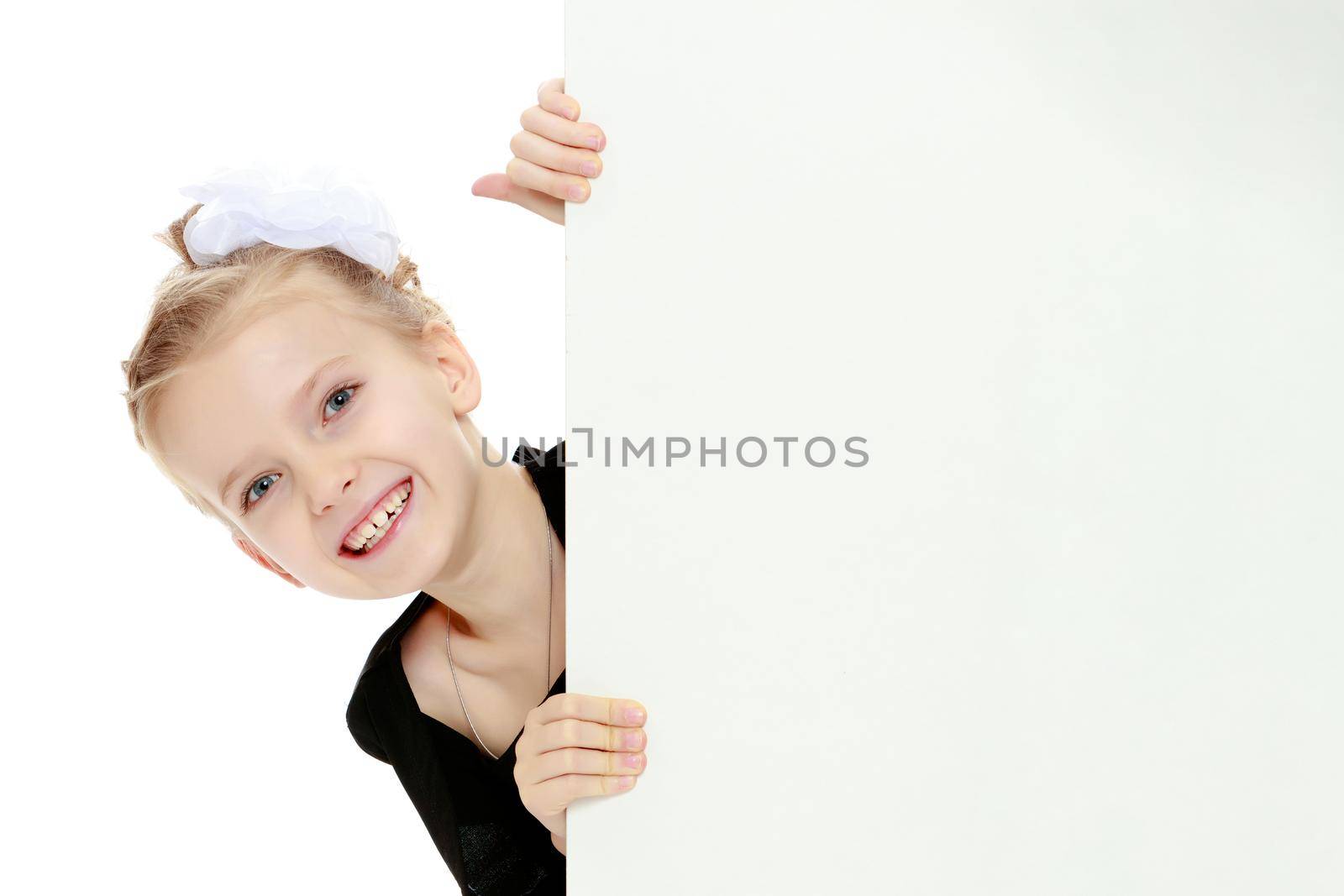 Beautiful little blonde girl dressed in a white short dress with black sleeves and a black belt.The girl peeks out from behind white banner.Close-up.Isolated on white background.