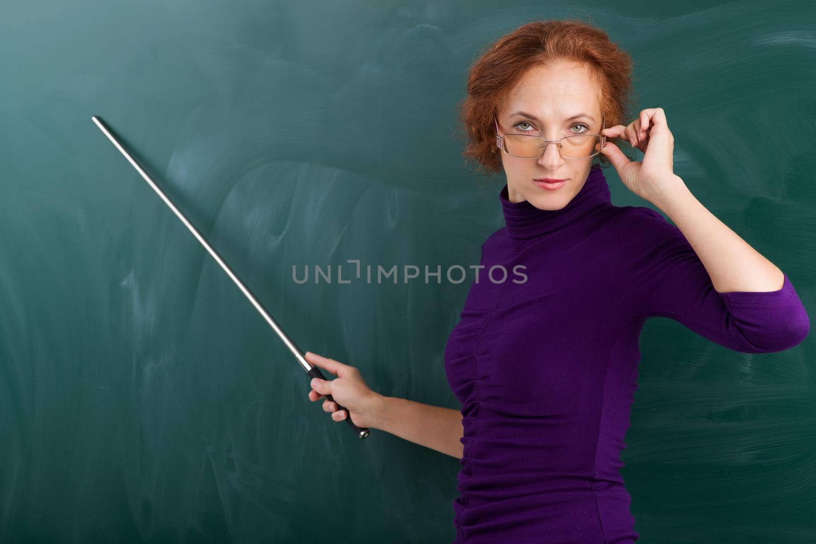 Teacher standing at blackboard with pointer. Portrait of smiling young woman looking over glasses. Teacher posing at green chalkboard and pointing at it